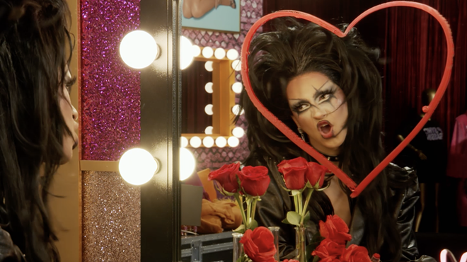 drag race looking at herself in the mirror. a heart is drawn in lipstick on the mirror