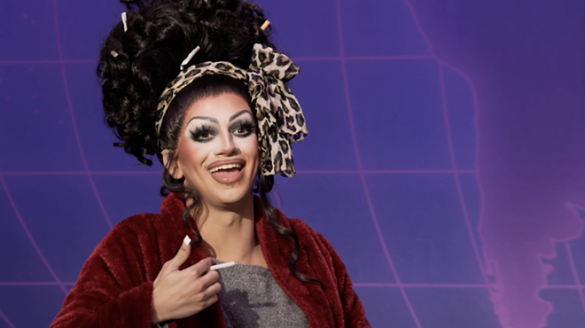 still image of an episode of RuPaul's drag race featuring Suzie Toot in drag makeup, a leopard-print headband, and holding a cigarette